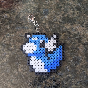 Op de afbeelding: Een blauwe en witte gepixelde sleutelhanger met een zilveren ketting. De sleutelhanger is in de vorm van een Pac-Man-personage.