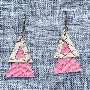 Puede incluir: Un par de pendientes de forma triangular con un diseño en capas de color rosa y blanco. Los pendientes tienen un patrón texturizado, similar a la piel de serpiente.