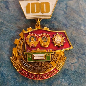 Puede incluir: Una medalla dorada con un diseño rojo, blanco y verde. La medalla presenta un engranaje con el texto "Кировский машиностроительный завод" e "им. В.И. Донина" alrededor del borde. La medalla también presenta una pancarta roja con el texto "1982" y una estrella con un martillo y una hoz.