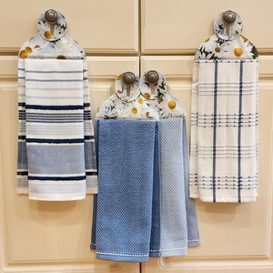 May include: Three kitchen towel holders with floral designs and blue and white striped towels hanging from them. The holders are attached to white cabinets.