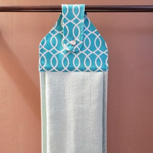 May include: A teal and white patterned fabric towel holder with a metal button. The holder is hanging on a brown bar and a light green towel is hanging from it.