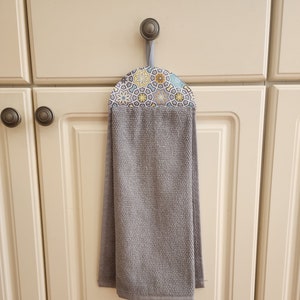 May include: A gray kitchen towel with a patterned fabric holder. The holder is attached to a cabinet door with a hook.