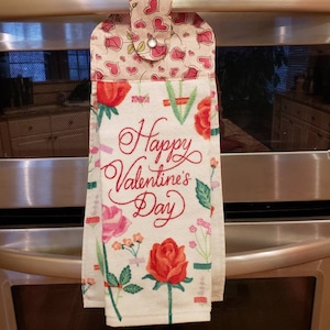 May include: A white kitchen towel with a floral pattern and the text "Happy Valentine's Day" in red. The towel has a pink and white fabric loop with a button closure.