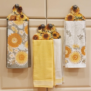 May include: Three kitchen towels with sunflower designs. The towels are hanging on hooks. One towel is yellow and white, one is gray and white, and one is white with a floral pattern.