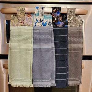 May include: Four kitchen towels hanging on a silver oven handle. The towels are made of a soft, absorbent fabric and have different patterns. The towels are green, blue, black, and brown.