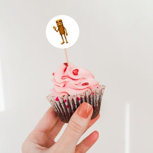 May include: A cupcake with pink frosting and heart-shaped sprinkles. A cartoon character on a white circle is attached to the top of the cupcake with a wooden stick. The cupcake is held in a person's hand.
