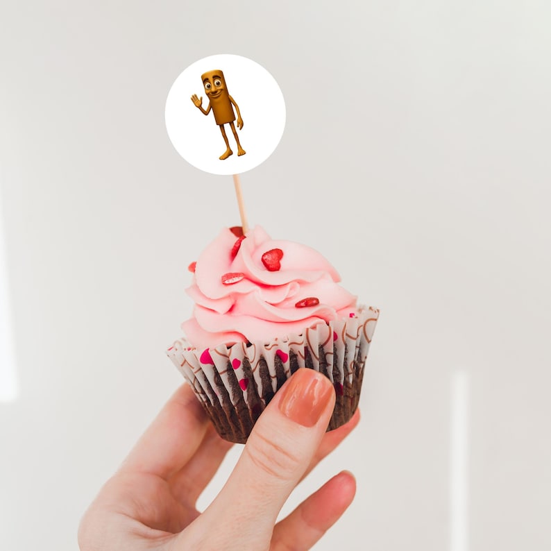 May include: A cupcake with pink frosting and heart-shaped sprinkles. A cartoon character on a white circle is attached to the top of the cupcake with a wooden stick. The cupcake is held in a person's hand.