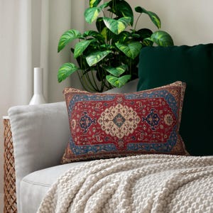 May include: A rectangular decorative pillow with a red, blue, and beige oriental rug design. The pillow is on a gray sofa with a white throw blanket. A green plant and a dark green pillow are in the background.