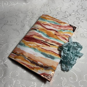 May include: A rectangular book cover with a colorful abstract pattern in shades of orange, red, and teal. The cover has decorative gold-colored corner accents. A small, crumpled, light blue paper decoration sits nearby.