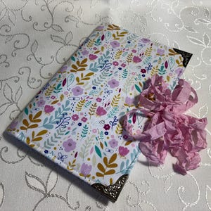 May include: A floral patterned journal with a white background, featuring pink, purple, and blue flowers, and gold leaves. The journal has decorative metal corner protectors and a pink ribbon. The journal is on a white floral patterned surface.