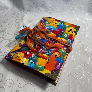 May include: A colorful fabric-covered journal with a bright orange, yellow, and blue pattern of cartoon dogs and cats. The journal is tied with a turquoise and orange ribbon.
