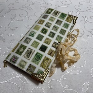 May include: A rectangular wallet with a white background covered in vintage postage stamp designs in green, brown, and cream colors. The corners are adorned with gold-colored metal accents. A cream-colored ribbon is tied to the side.