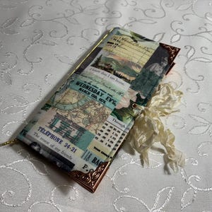 May include: A rectangular journal with a vintage collage design featuring maps, botanical illustrations, and text fragments. The journal has a gold-colored metal frame and a cream-colored ribbon. The text on the journal reads "WEDNESDAY EV'G, MARCH 15th, 1876" and "TELEPHONE 24-21".