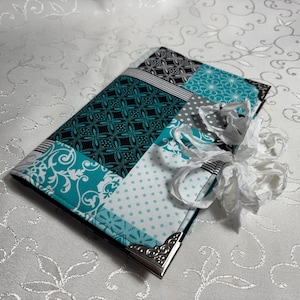 Op de afbeelding: Een rechthoekig notitieboek met een patchwork-ontwerp. De omslag heeft stoffen met patronen in turquoise, zwart, wit en grijs. Een wit lint en een strik zijn om het notitieboek gebonden. Zilveren hoekbeschermers zijn zichtbaar.