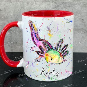 Pu&ograve; includere: Una tazza in ceramica bianca con un bordo e un manico rossi. La tazza presenta un'illustrazione ad acquerello colorata di un axolotl con il nome "Karly" scritto sotto.