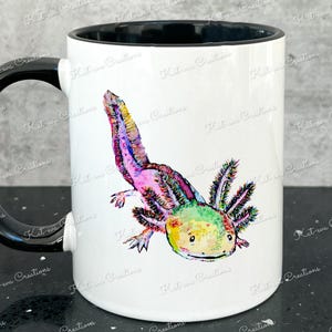 Pu&ograve; includere: Una tazza in ceramica bianca con un manico nero e un bordo nero. La tazza presenta un'illustrazione colorata di un axolotl, un tipo di salamandra, con colori rosa, verde e giallo.