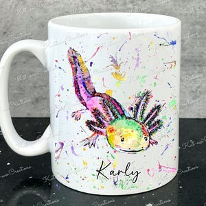 Pu&ograve; includere: Una tazza in ceramica bianca con un'illustrazione ad acquerello colorata di un axolotl. L'axolotl &egrave; rosa, verde e giallo con un contorno nero. Lo sfondo &egrave; bianco con schizzi di vernice colorata. La tazza ha un manico nero e il nome "Karly" &egrave; scritto in nero sotto l'axolotl.