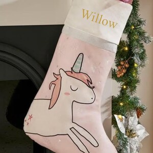 May include: Pink Christmas stocking featuring a unicorn design. The stocking has a white cuff with the name "Willow" in gold lettering. The unicorn is white with a pink mane and a blue horn. The stocking is filled with wrapped gifts.