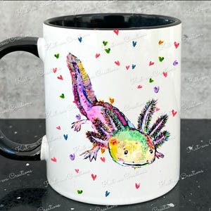 Pu&ograve; includere: Una tazza in ceramica bianca con manico nero presenta un'illustrazione colorata di un axolotl con tonalit&agrave; rosa, verdi e gialle. L'axolotl &egrave; circondato da piccoli cuori colorati.