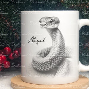 May include: White ceramic mug featuring a black and white illustration of a snake. The name "Abigail" is written in cursive on the mug.