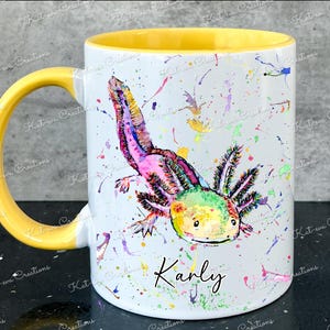 Pu&ograve; includere: Una tazza in ceramica bianca con un bordo e un manico gialli. La tazza presenta un'illustrazione ad acquerello colorata di un axolotl con il nome "Karly" scritto sotto.
