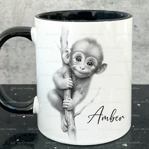 May include: A white ceramic mug with a black handle and black interior. The mug features a black and white illustration of a monkey holding onto a branch. The name "Amber" is written in black cursive at the bottom of the mug.