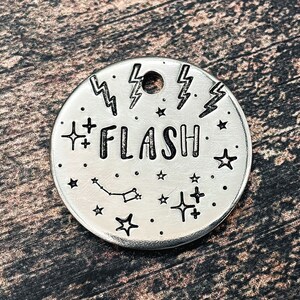 Peut inclure: Une médaille de chien en argent avec un motif noir et blanc représentant des étoiles, des éclairs et le mot "FLASH".