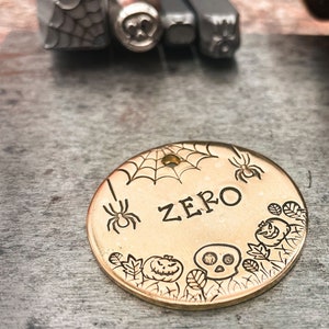May include: Gold-colored metal dog tag with a spider web, spiders, pumpkins, and a skull design. The tag says "ZERO" in a black font.