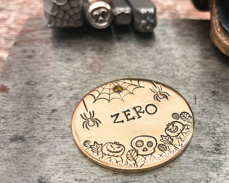 May include: Gold-colored metal dog tag with a spider web, spiders, pumpkins, and a skull design. The tag says "ZERO" in a black font.