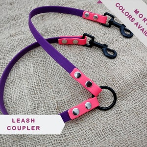 Op de afbeelding: Een paarse en roze hondenriemkoppeling met zwarte metalen sluitingen en een zwarte metalen ring. De hondenriemkoppeling heeft de tekst "LEASH COUPLER" en "MORE COLORS AVAILABLE".