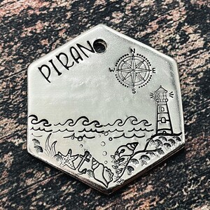 Könnte beinhalten: Eine silberfarbene, sechseckige Haustier-Marke mit dem Wort "PIRANO" oben. Die Marke zeigt eine Windrose, einen Leuchtturm, Wellen und Illustrationen des Meereslebens. Oben befindet sich ein Loch zur Befestigung.