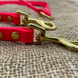 May include: A bright pink dog lead with two gold metal clasps. The lead is lying on a brown hessian surface.