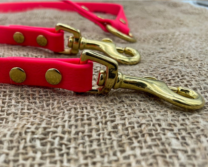 May include: A bright pink dog lead with two gold metal clasps. The lead is lying on a brown hessian surface.