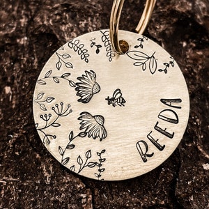 Peut inclure: Une étiquette en métal doré avec un motif floral et le nom "RIEDA" gravé dessus. L'étiquette est attachée à un anneau en métal doré.