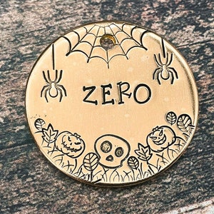 May include: Gold-colored metal pet tag with a spider web design, two spiders, a skull, and two pumpkins. The tag is engraved with the name "ZERO".