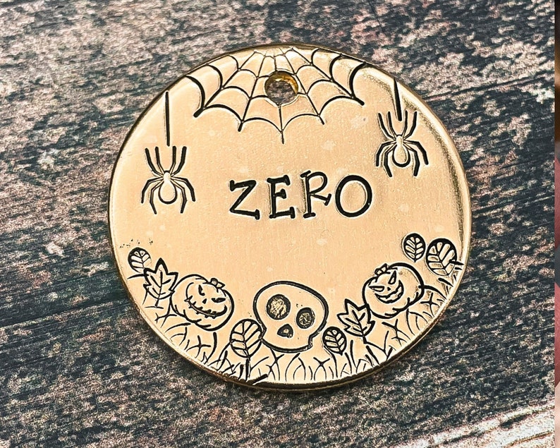 May include: Gold-colored metal pet tag with a spider web design, two spiders, a skull, and two pumpkins. The tag is engraved with the name "ZERO".