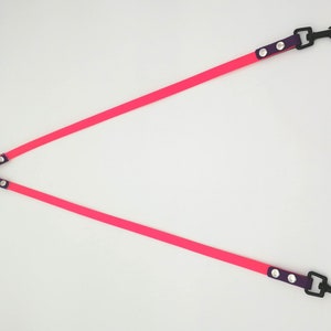 May include: A bright pink, Y-shaped dog lead with black metal clips and a black metal ring. The lead has purple accents with silver rivets. The lead is designed for walking two dogs at once.