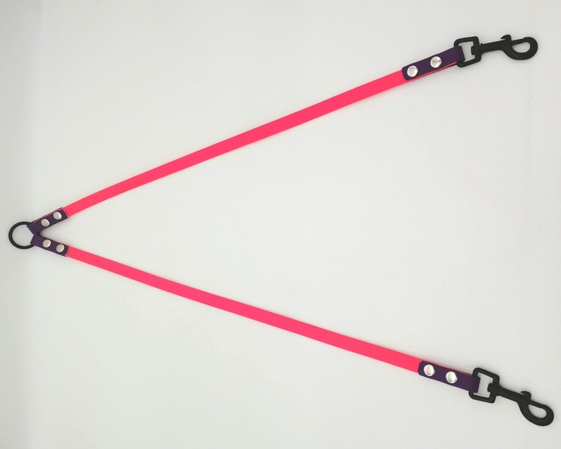 May include: A bright pink, Y-shaped dog lead with black metal clips and a black metal ring. The lead has purple accents with silver rivets. The lead is designed for walking two dogs at once.