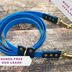 Puede incluir: Una correa para perros manos libres azul con dos clips metálicos y detalles negros. La correa está enrollada sobre una superficie de madera. "Más colores disponibles" y "Correa para perros manos libres" están escritos en blanco sobre un fondo morado.