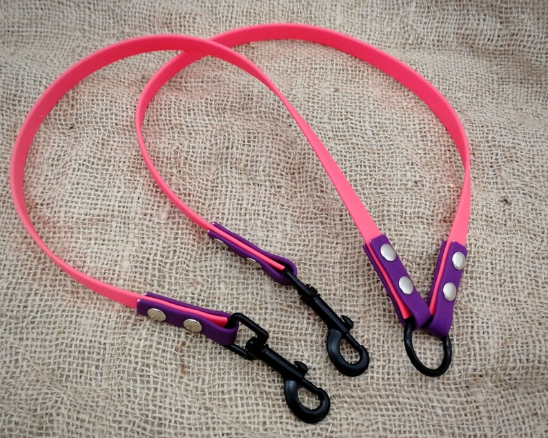 May include: Two neon pink dog leads with purple and pink detailing. Each lead has a black metal clip and is attached to a purple and pink handle with silver rivets. The leads are shown on a textured, beige surface.