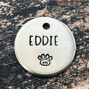 Op de afbeelding: Een ronde, zilveren dierenpenning met de naam "EDDIE" in zwarte letters. Onder de naam staat een pootafdruk met een hart. De penning heeft een gat aan de bovenkant om te bevestigen.