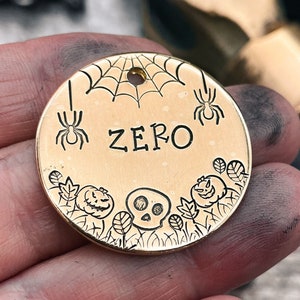 May include: Gold-colored metal dog tag with a Halloween theme. The tag features a spider web, spiders, a skull, pumpkins, and leaves. The name "ZERO" is engraved on the tag.