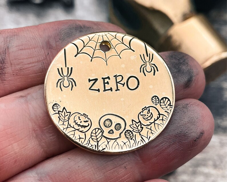 May include: Gold-colored metal dog tag with a Halloween theme. The tag features a spider web, spiders, a skull, pumpkins, and leaves. The name "ZERO" is engraved on the tag.