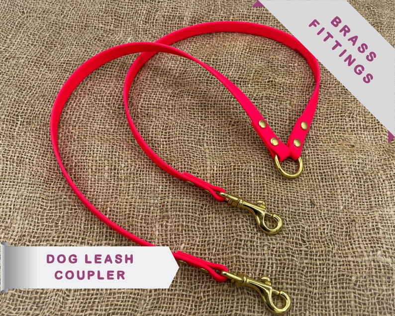 May include: A pink dog leash coupler with two brass swivel clips and a brass D-ring. The leash is made of durable, flexible material.  "BRASS FITTINGS" and "DOG LEASH COUPLER" are printed on the image.
