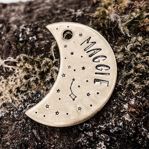 May include: A brass crescent moon pet tag with a constellation design and the name "Maggie" engraved on it.