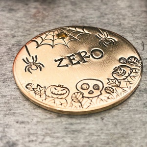 May include: Gold-colored metal tag with a spider web, spider, skull, and pumpkin design. The tag is engraved with the word "ZERO".