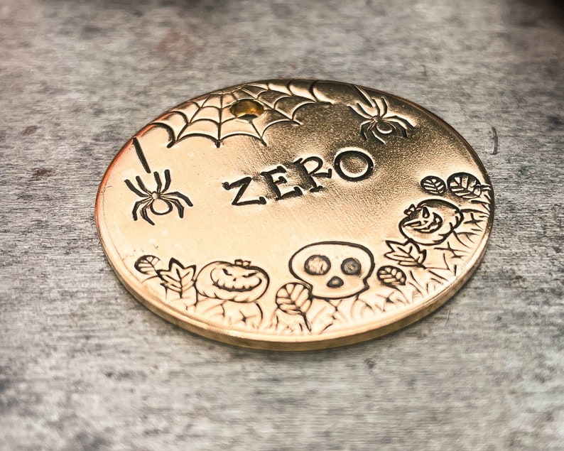 May include: Gold-colored metal tag with a spider web, spider, skull, and pumpkin design. The tag is engraved with the word "ZERO".