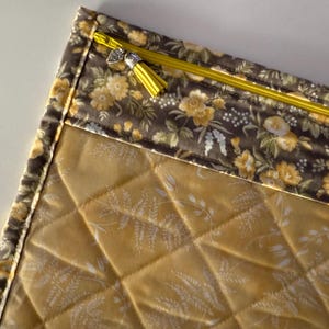 Könnte beinhalten: Eine gesteppte Reißverschlusstasche mit einem Blumendruck auf der oberen Klappe und einem einfarbigen, gesteppten gelben Stoff auf der Unterseite. Der Reißverschluss hat einen gelben Quasten-Zipper.