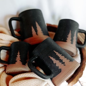 May include: Four black ceramic mugs with a brown glaze and a silhouette of pine trees. The mugs are stacked on top of each other and are sitting on a wooden surface.