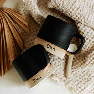 May include: Two black ceramic mugs with a tan base and the word "Dad" printed on them. The mugs have a simple, modern design with a handle. The mugs are on a neutral-toned surface.